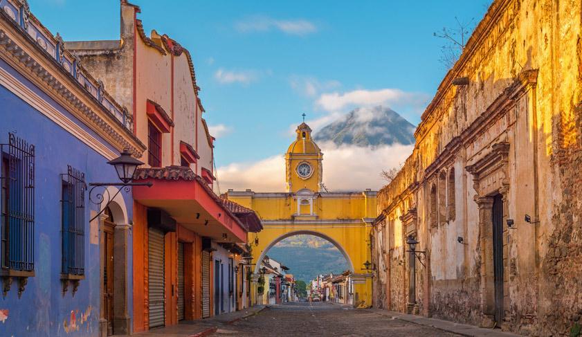 The colorful main street of Antigua City at sunrise with the famous yellow arch in the center and the active Agua volcano